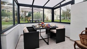A conservatory with a dining table set for breakfast, surrounded by wicker chairs, large windows, and a view of the garden. - Le Gîte de Gravelodic (PAIMPOL)
