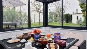 A breakfast table set with various food items and drinks, a vase with flowers, and a view of the garden through large windows. - Le Gîte de Gravelodic (PAIMPOL)