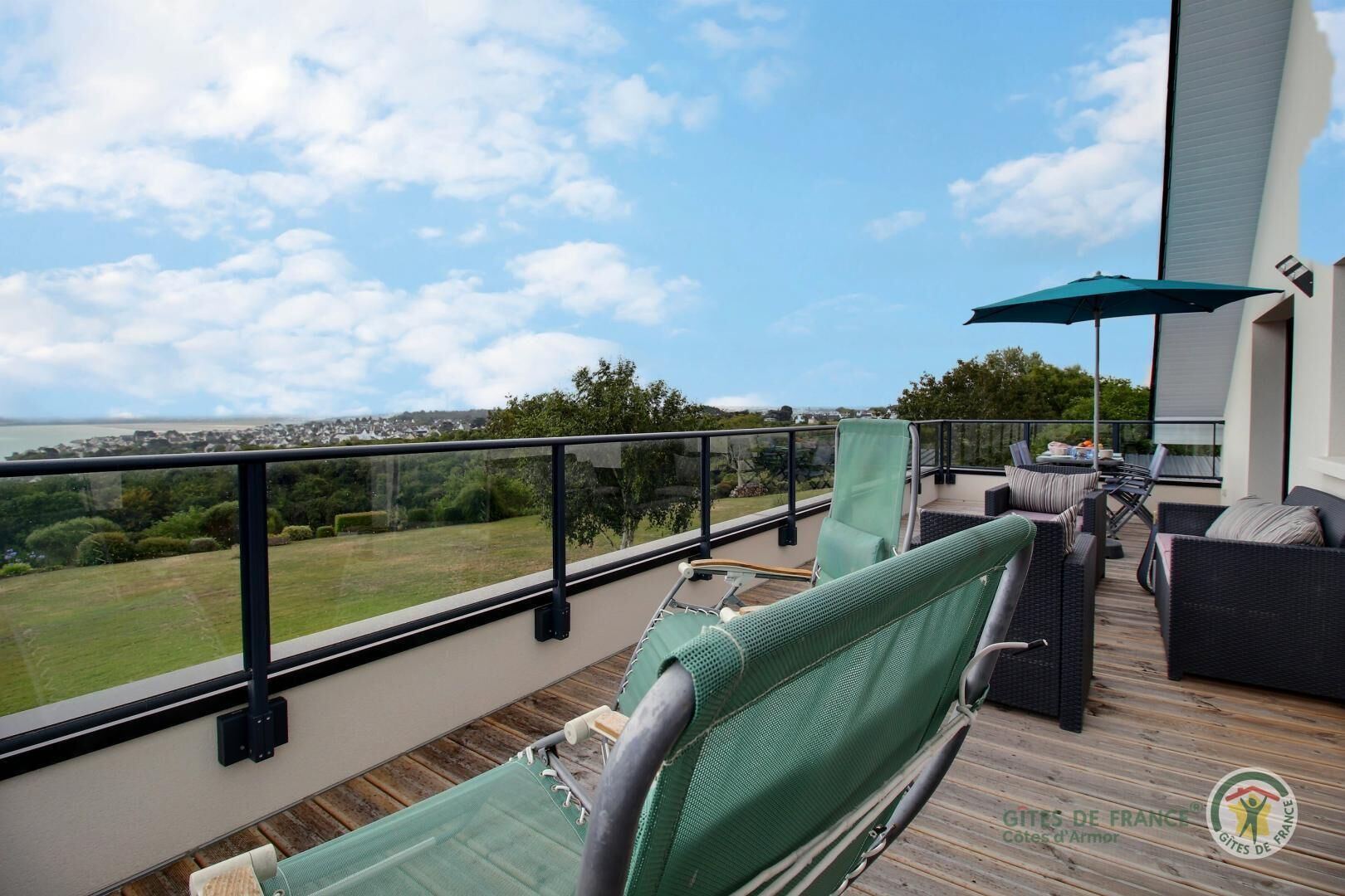 Een terras buiten met rieten meubilair, een groene parasol en uitzicht op een kustlandschap.