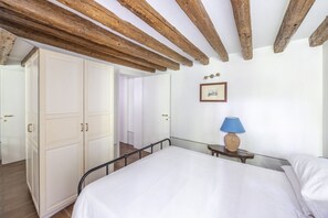 A bedroom with a bed, wooden ceiling beams, a wardrobe, and a bedside table with a lamp.