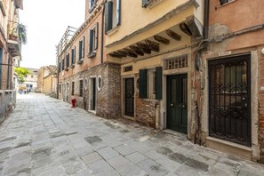 A narrow cobblestone street lined with historic buildings, featuring a mix of brick and plaster facades, traditional shutters, and ornate metal gates.