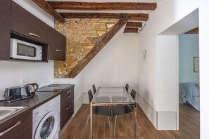 A compact kitchen with a built-in microwave, toaster, and washing machine. A glass dining table with chairs is positioned under a sloped ceiling with exposed wooden beams. - Ca' Gamba, a quite typical space in Venice (Venezia)