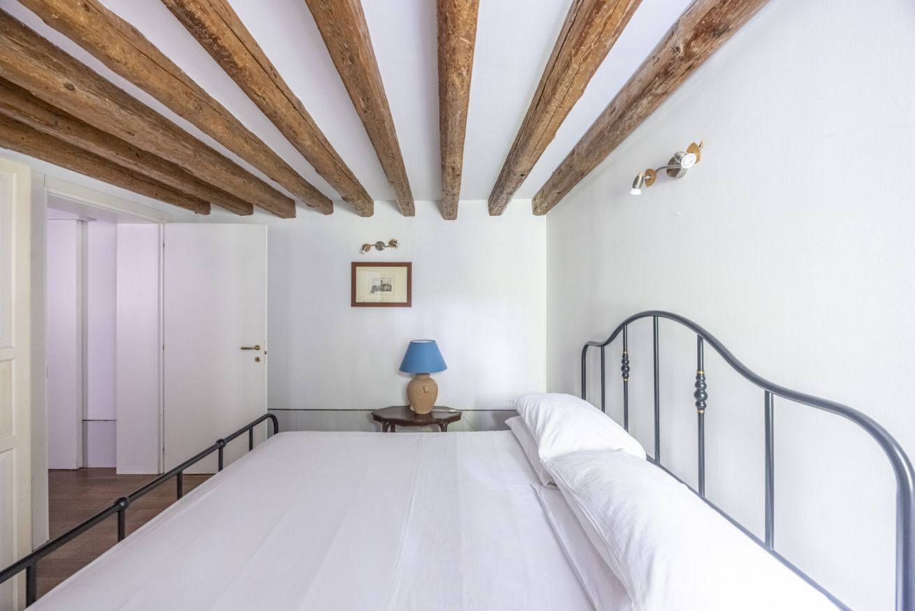 Un dormitorio con techo de madera, una cama con ropa de cama blanca, una mesita de noche pequeña con una lámpara y un cuadro enmarcado en la pared.