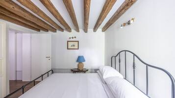 Un dormitorio con techo de madera, una cama con ropa de cama blanca, una mesita de noche pequeña con una lámpara y un cuadro enmarcado en la pared.