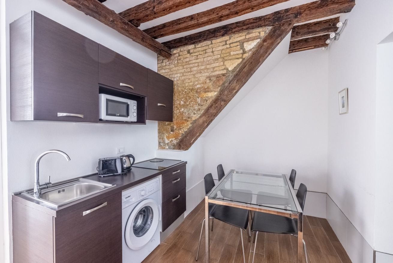 A compact kitchen with a washing machine, a microwave, and a toaster. There is a glass table with chairs and a brick wall in the background.