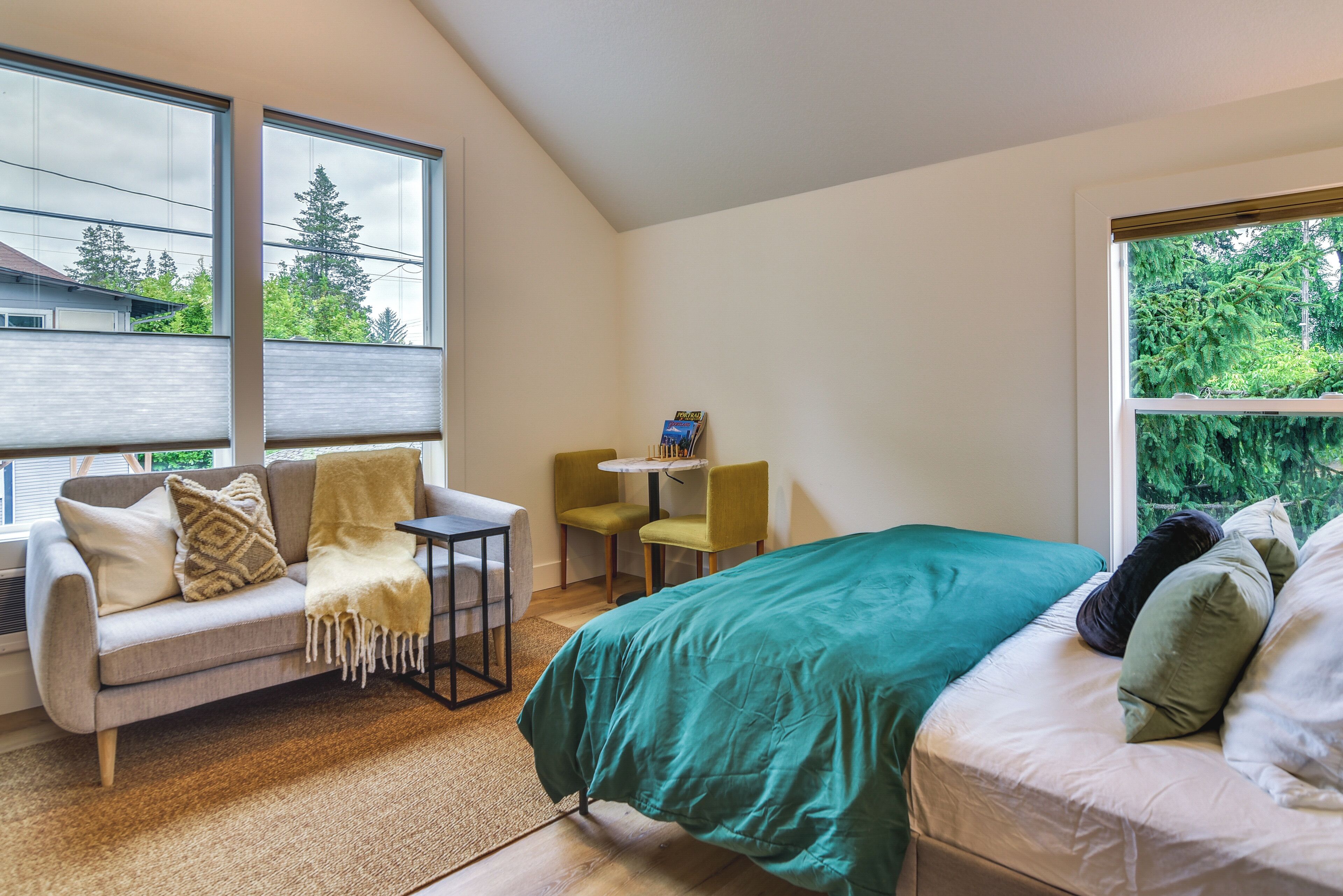 A bedroom with a bed, a sofa, a small table, and two chairs. There are large windows providing a view of trees and a house.