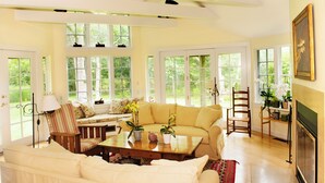 A spacious living room with a large sofa, a coffee table, and a fireplace. There are multiple windows providing natural light and views of the outdoors. - Spacious and gracious architect designed home on 3 acres off Middle Road. (Chilmark)