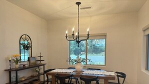 Un comedor con una mesa de madera, seis sillas, una araña, un ventanal con vistas, un espejo y una estantería con objetos decorativos.