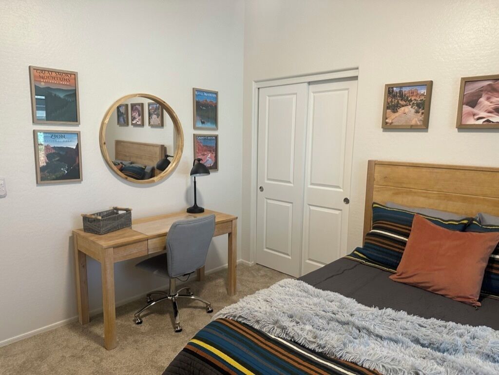 Een slaapkamer met een houten bureau, een stoel, een bed met gestreepte lakens en een spiegel aan de muur.