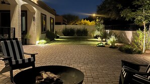 Un patio al aire libre con un hogar para fuego, dos sillones y un sendero de ladrillos que conduce a un edificio con puertas arqueadas.