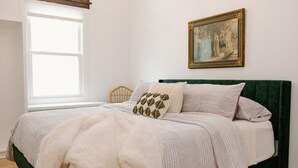 Une chambre à coucher avec un lit, une fenêtre, un tableau et une tête de lit recouverte d’un tissu vert.
