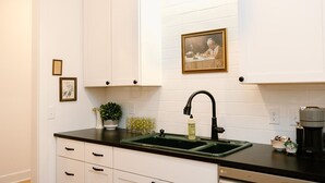 Une cuisine avec des meubles blancs, un plan de travail noir, un évier vert, une machine à café et un tableau encadré au mur.