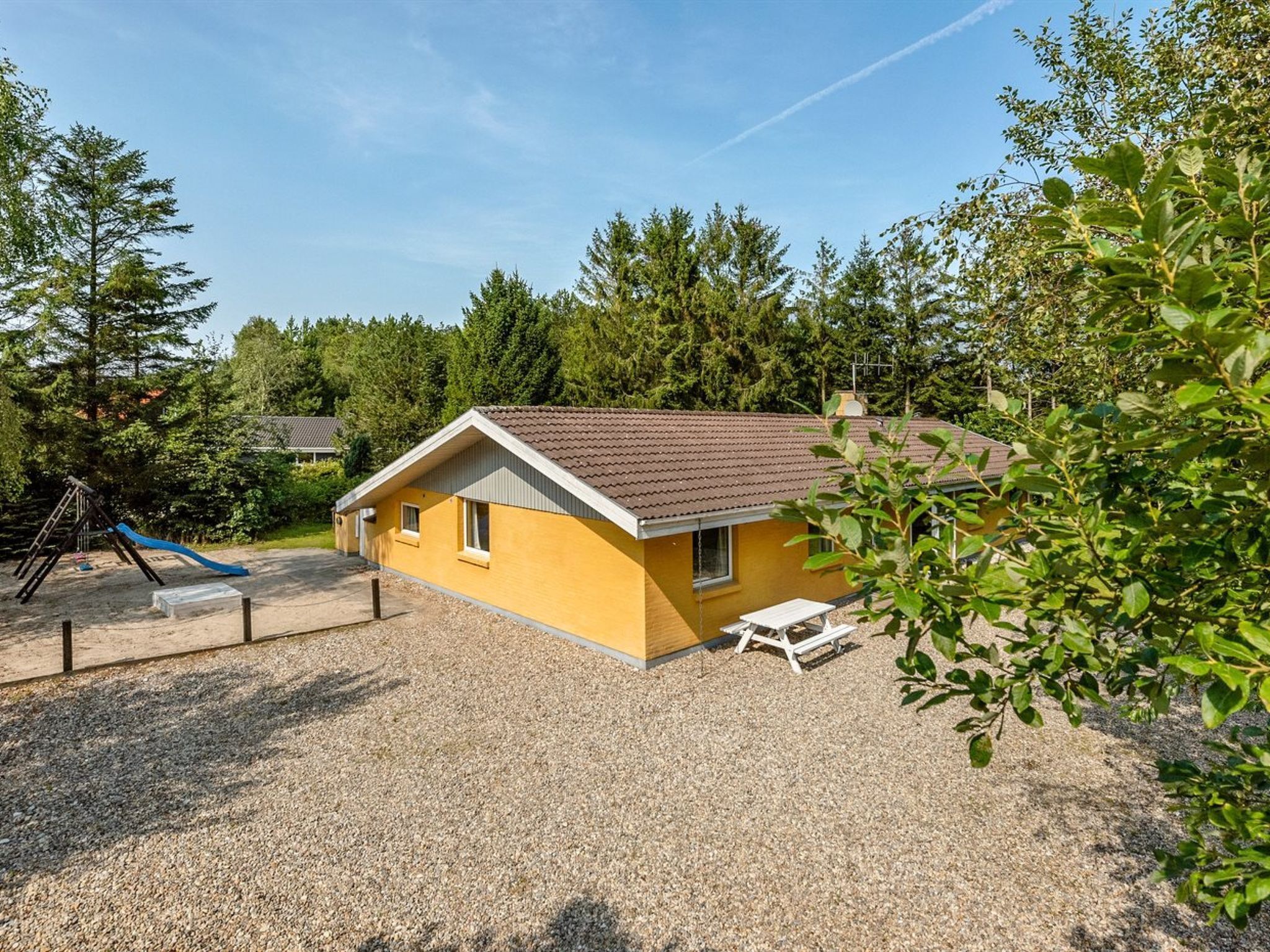 A yellow building with a brown roof, surrounded by gravel and trees.