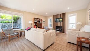 A spacious living room with a large sofa, a smaller sofa, and a chair. There is a bookshelf, a television mounted on the wall, and a fireplace. - WestCoast Garden Suite | 1 Bedroom | Private | Quiet | Central | Modern (North Vancouver)