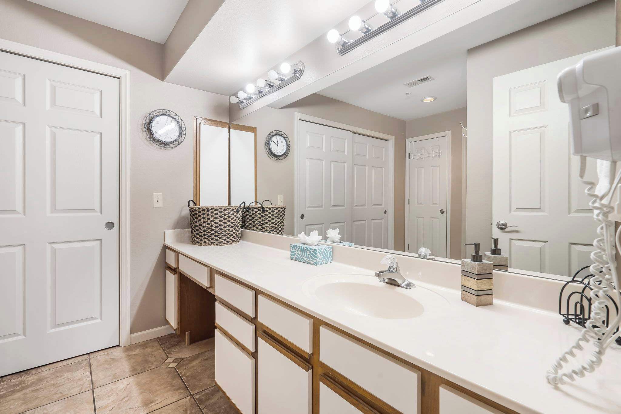 A bathroom with a white vanity, a large mirror, and a sink. There are two clocks on the wall, a towel rack, and a hairdryer on the counter.