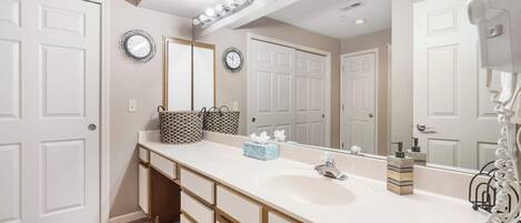 A bathroom with a white vanity, a large mirror, and a sink. There are two clocks on the wall, a towel rack, and a hairdryer on the counter.