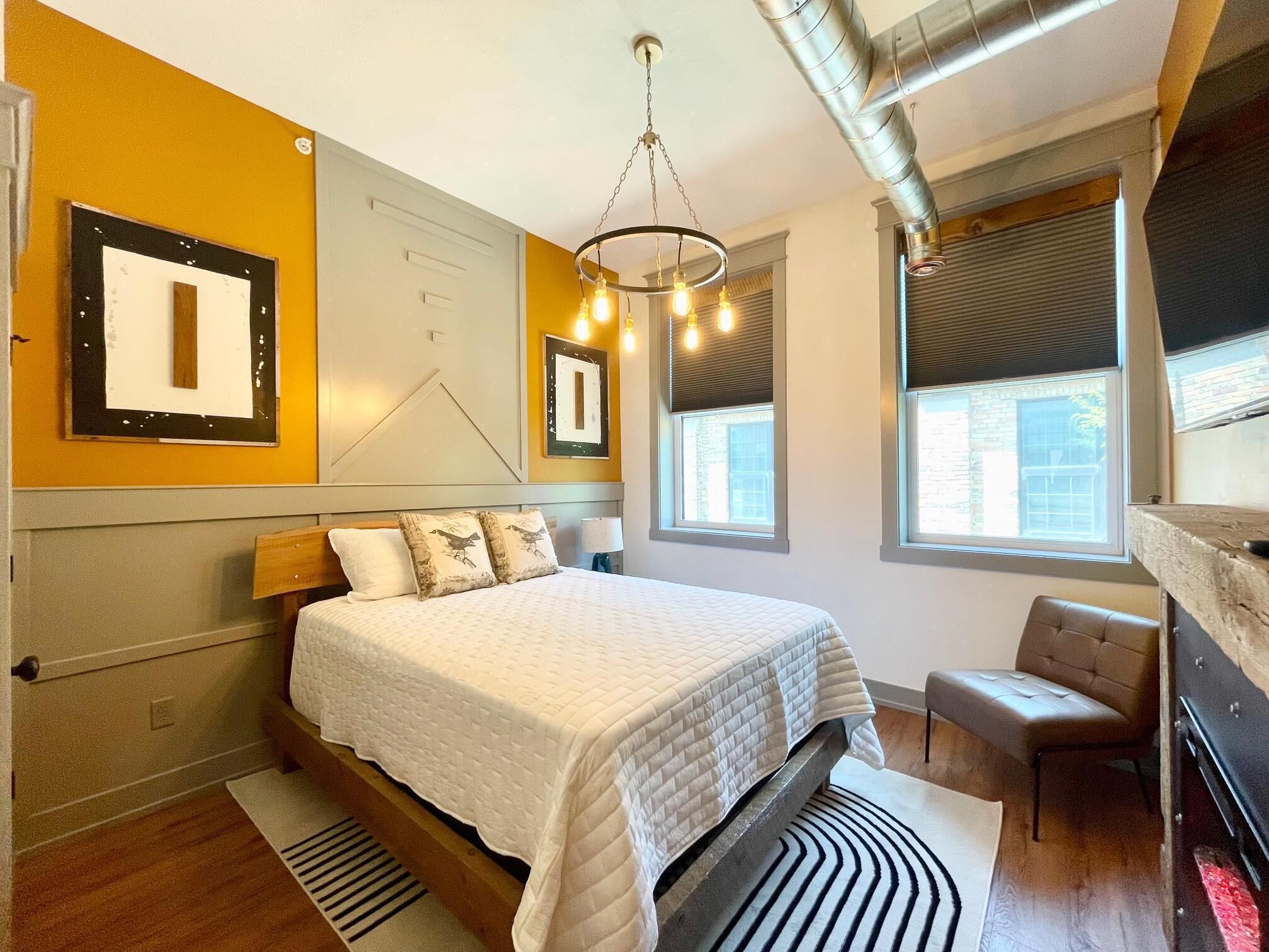 Une chambre moderne avec un grand lit, un bureau et une chaise. On y trouve des fenêtres équipées de stores, un lustre et une cheminée.