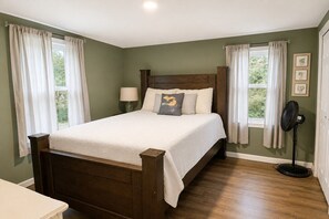 A bedroom with a wooden bed, two windows with curtains, a bedside table, a lamp, and a standing fan.