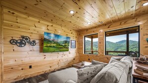 L’intérieur d’un chalet en bois chaleureux et accueillant, avec un canapé, un tableau représentant un paysage et une fenêtre donnant sur des collines verdoyantes.