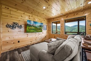 L’intérieur d’un chalet en bois chaleureux et accueillant, avec un canapé, un tableau représentant un paysage et une fenêtre donnant sur des collines verdoyantes.