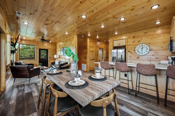A wooden interior with a dining table set for four, a kitchen area, and a large wall clock.