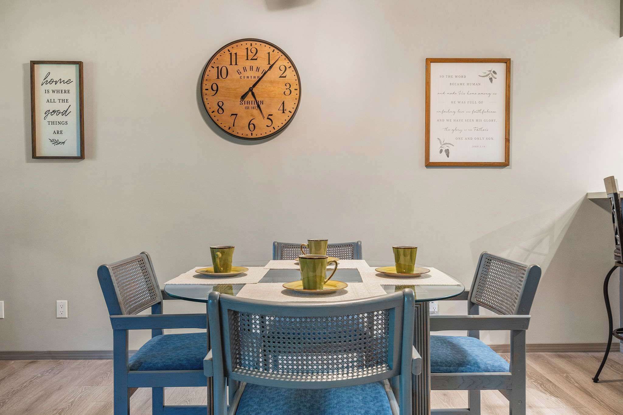 Een eethoek met een ronde wandklok, raamdecoratie met inspirerende citaten, en een tafel voor vier personen met blauwe stoelen.