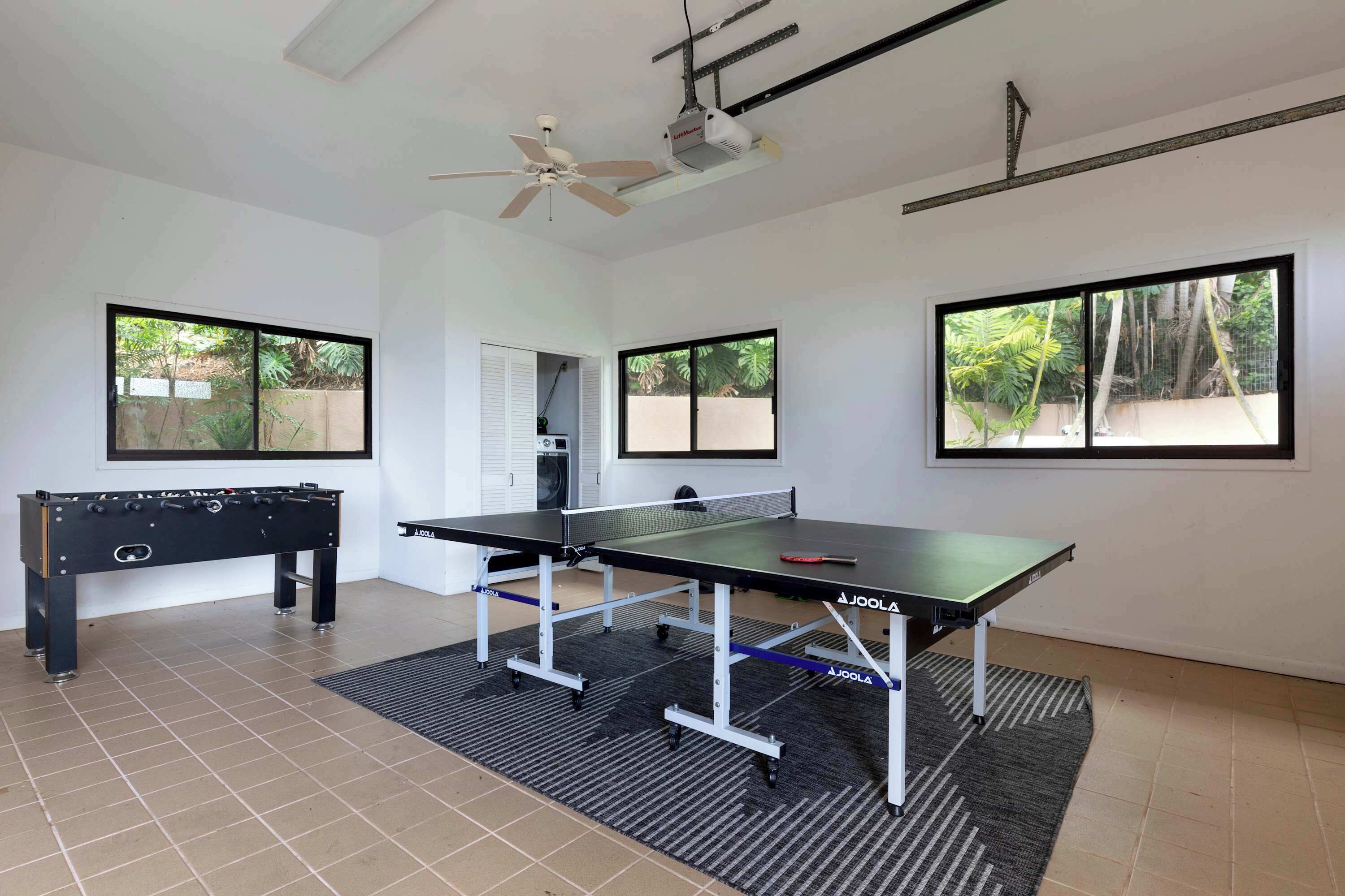 Quarto com mesa de pebolim, mesa de pingue-pongue, ventilador de teto e grandes janelas com vista para o verde.