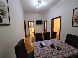 A dining area with a table set for four, a television mounted on the wall, and a kitchen visible through an open door.