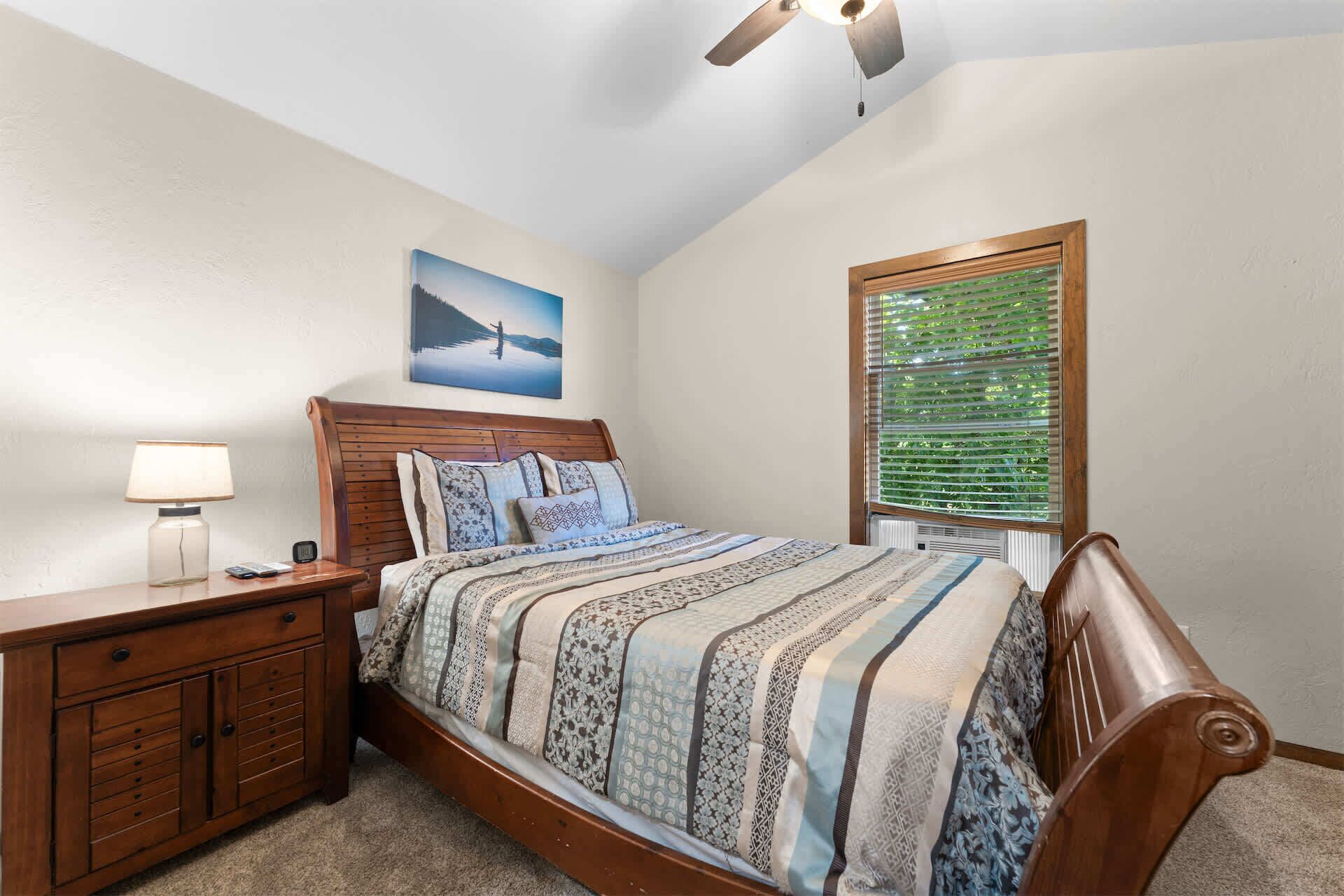 A bedroom with a wooden bed, a nightstand, a lamp, a painting, and a window with blinds.