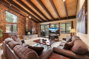 Amplia sala de estar con chimenea de piedra, sofás de cuero, techo de madera y ventanas grandes que ofrecen vistas al exterior.