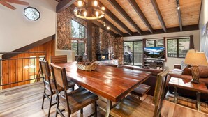 A spacious living area with a large wooden dining table, comfortable seating, a stone fireplace, and large windows offering views of the outdoors. - Heavenly Haven - Hot Tub + Seasonal HOA Pool (Zephyr Cove)