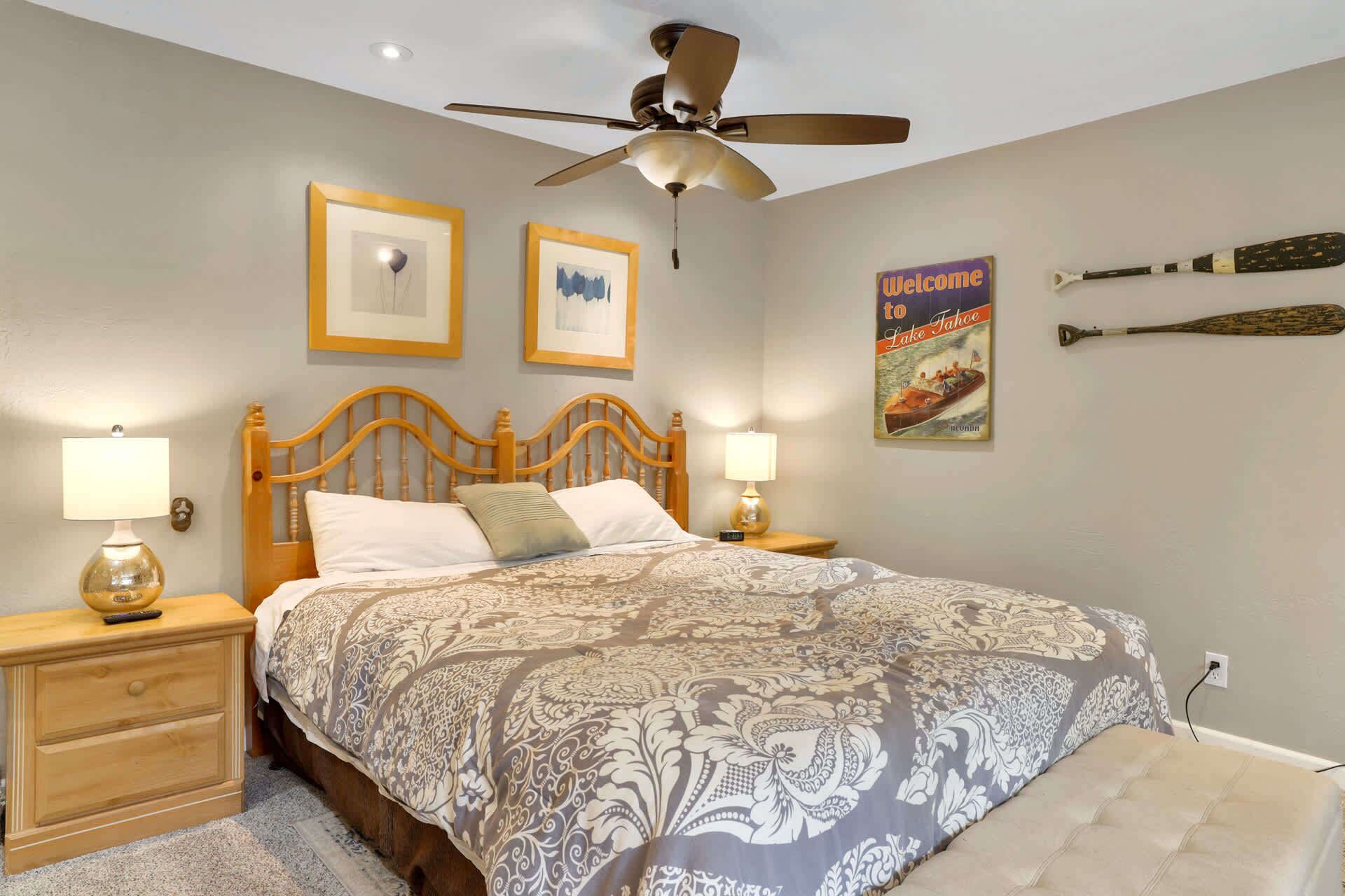 A bedroom with a wooden bed, two bedside tables with lamps, a ceiling fan, framed pictures, and a welcome sign.