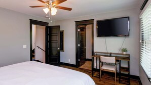 Un dormitorio con cama, escritorio y silla, ventilador de techo y televisor en la pared.