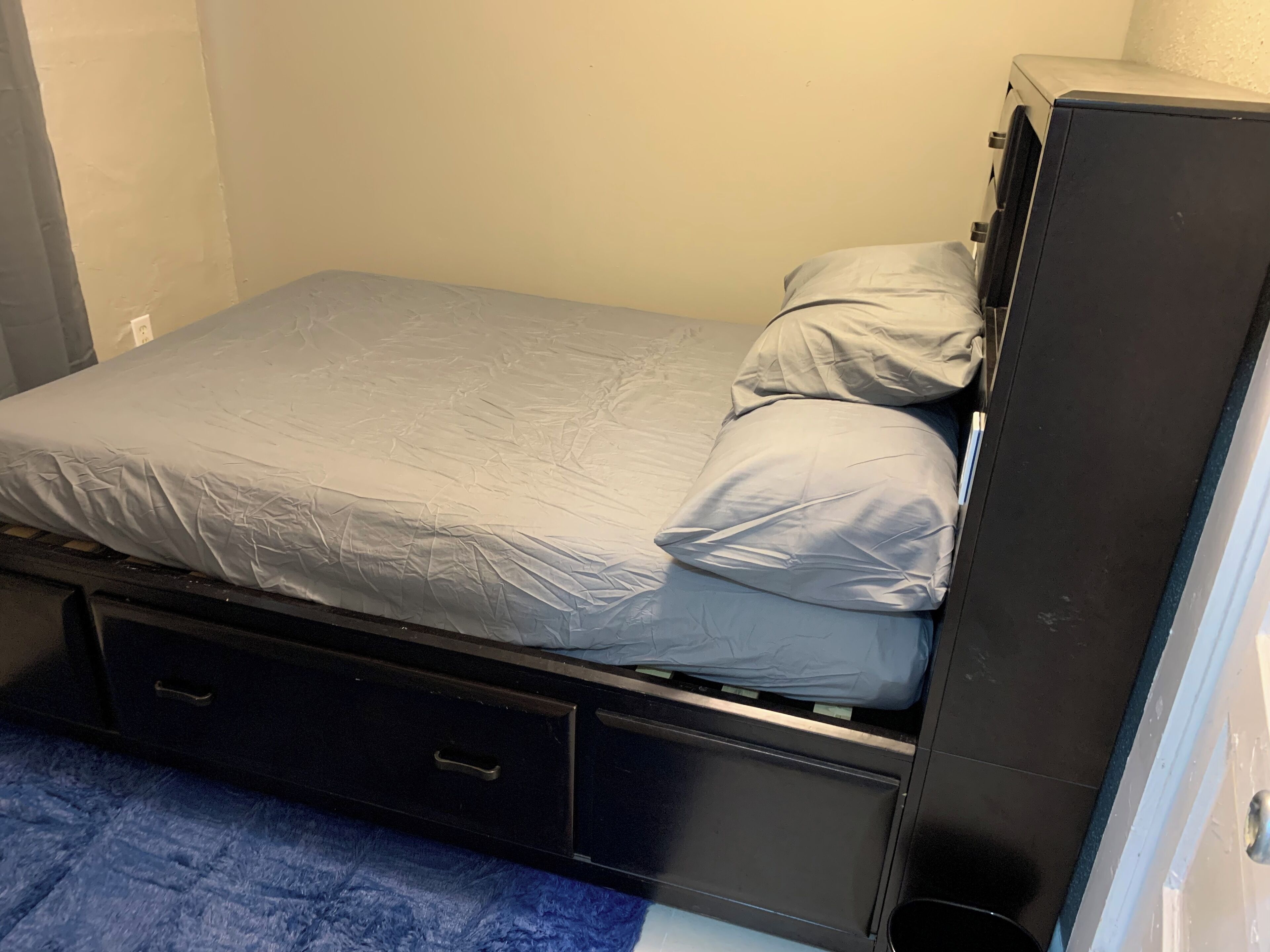 A small, single-bed room with a grey mattress, two pillows, a small drawer unit, and a blue carpet.