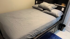 Ein Einzelbett mit grauem Matratze und Kissen, ein schwarzer Kopfseitenrahmen mit Schubladen und ein blauer Teppich auf dem Boden.