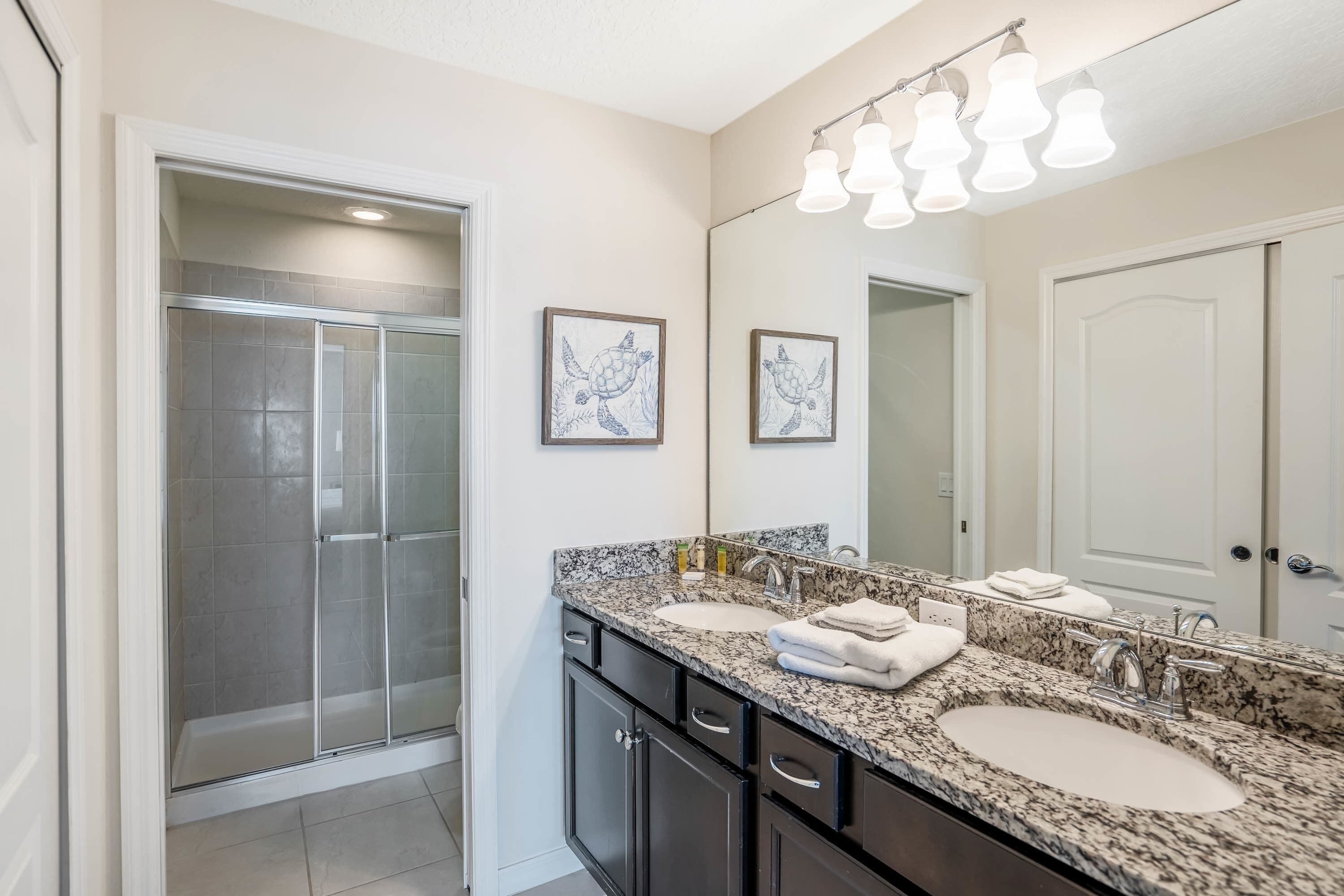 A bathroom with a glass shower, a double sink vanity, and two framed artwork on the wall.