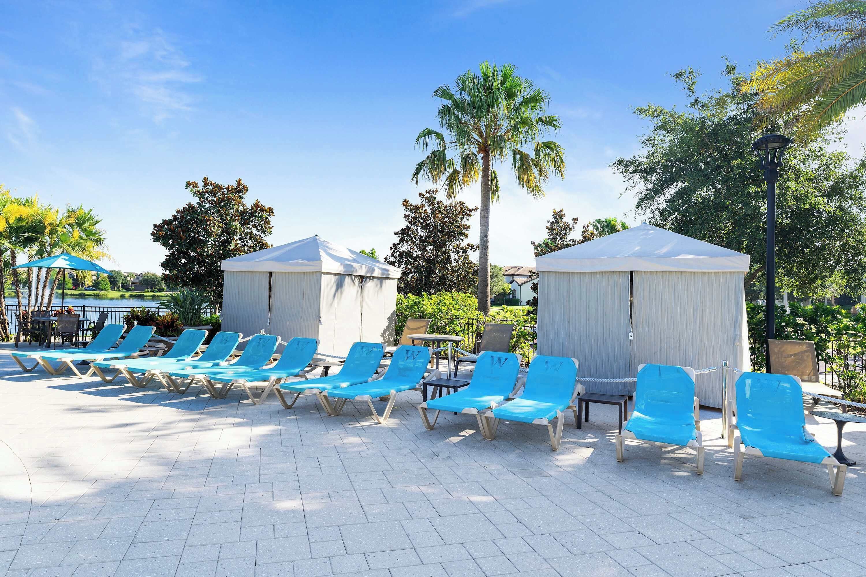 A poolside area with blue lounge chairs and white canopies, surrounded by palm trees and greenery.