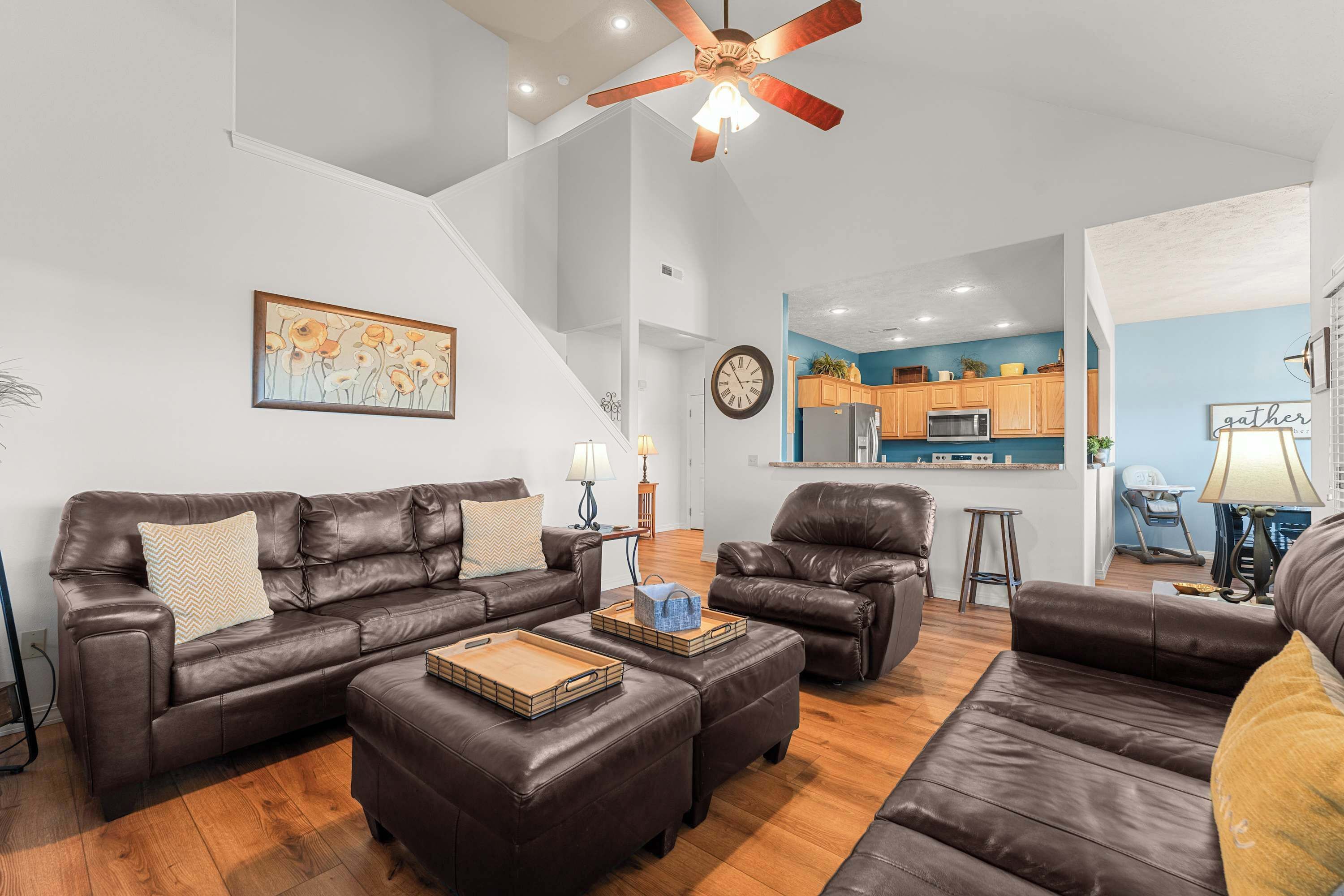 A spacious living room with a brown leather sofa set, wooden ottomans, and a ceiling fan. A kitchen is visible in the background with wooden cabinets and a blue accent wall.