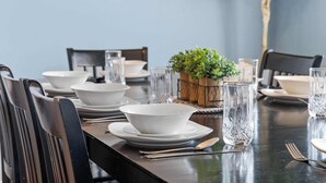 Uma mesa de jantar posta com tigelas, pratos e talheres brancos, além de copos transparentes e um vaso com planta.