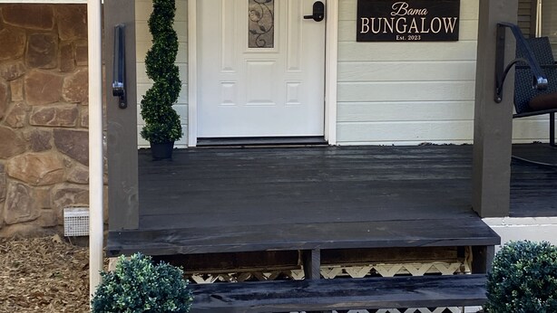 Ein Eingangsbereich mit einer weißen Tür, einem Willkommensschild, einem Schild mit der Aufschrift 'Bama Bungalow Est. 2023' und einer kleinen Veranda mit Stufen und Blumentöpfen.