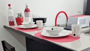 Un pequeño espacio de cocina con microondas, un grifo rojo y una encimera blanca. Hay dos sillas y una mesa pequeña con un mantel individual rojo.
