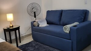 Una sala de estar con un sofá azul, una mesita redonda con una lámpara, un ventilador de pie y un cuadro en la pared.