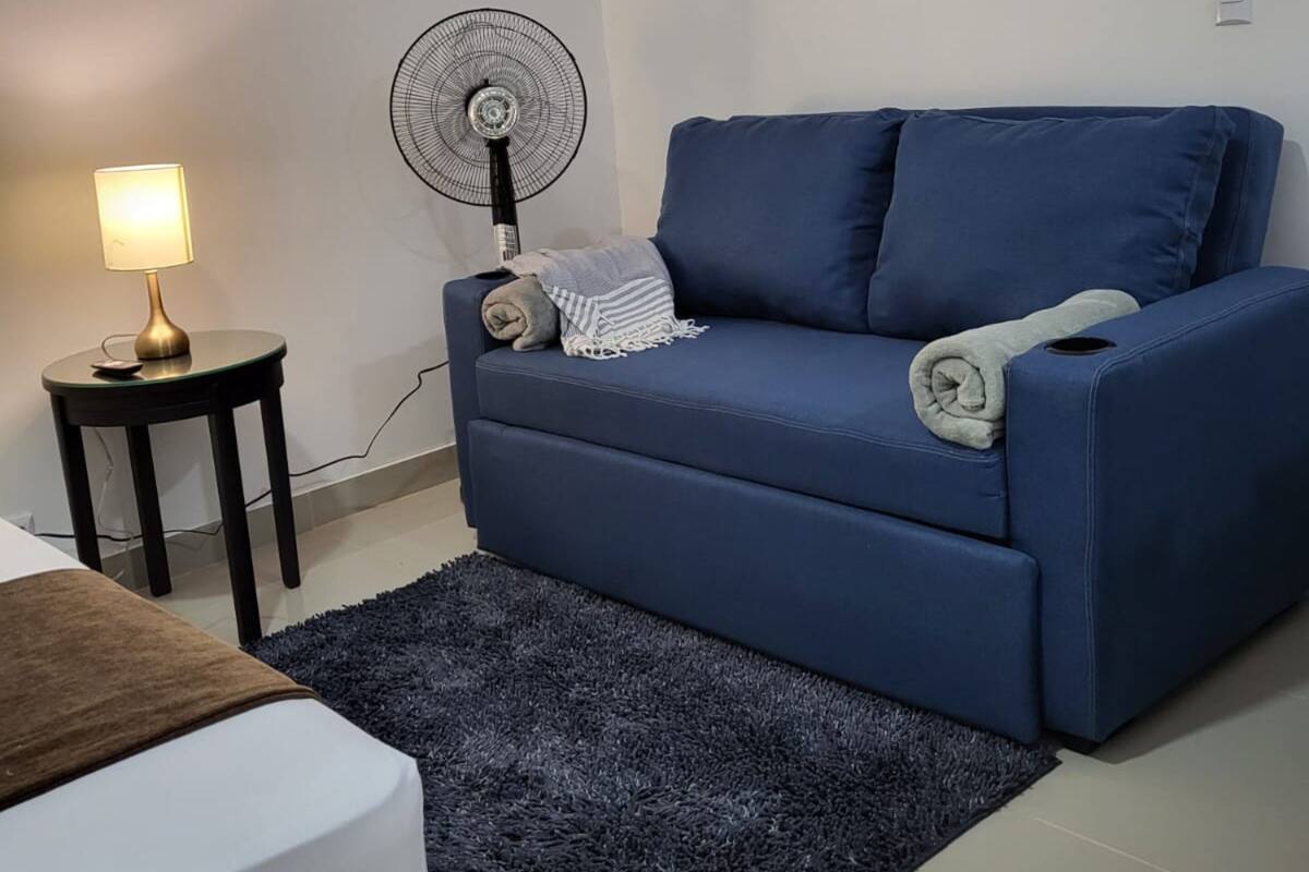 A living room with a blue sofa, a standing fan, a lamp, and a framed picture on the wall.