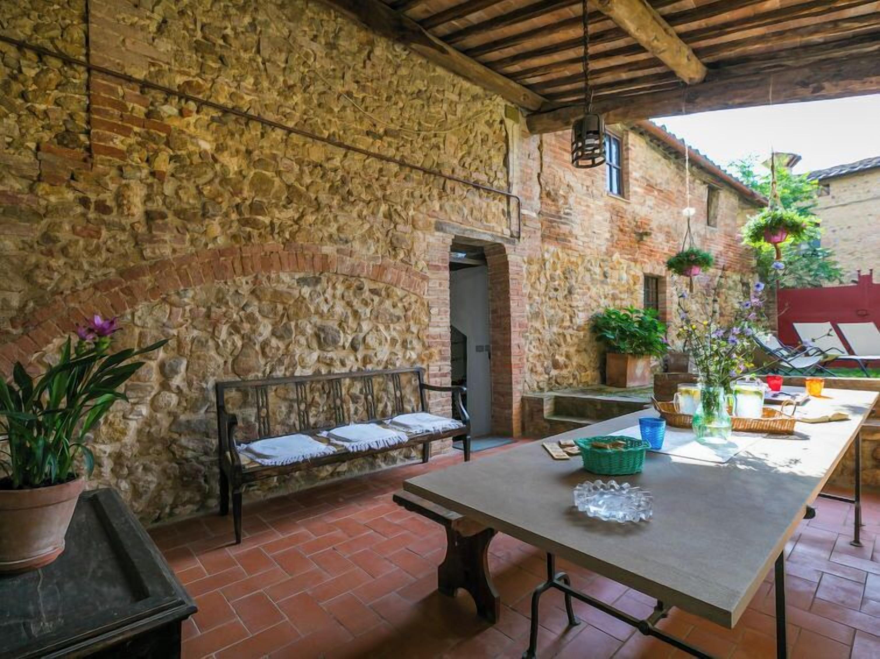 An outdoor patio with a stone wall, a wooden bench, a table with various items, and hanging plants.