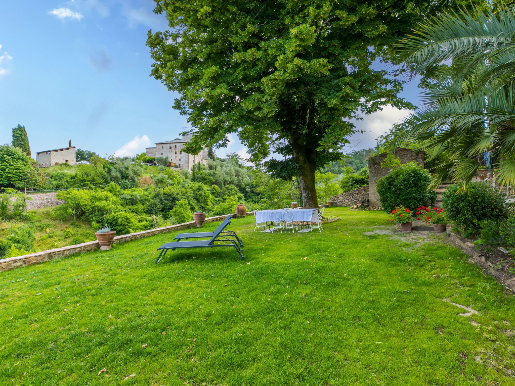 Un jardin luxuriant et verdoyant, avec une table et des chaises disposées pour les repas, entouré d’arbres et d’un mur de pierre.