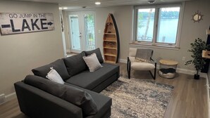 A living room with a dark grey sofa, a small white chair, a wooden bookshelf, and a sign that reads "GO JUMP IN THE LAKE".