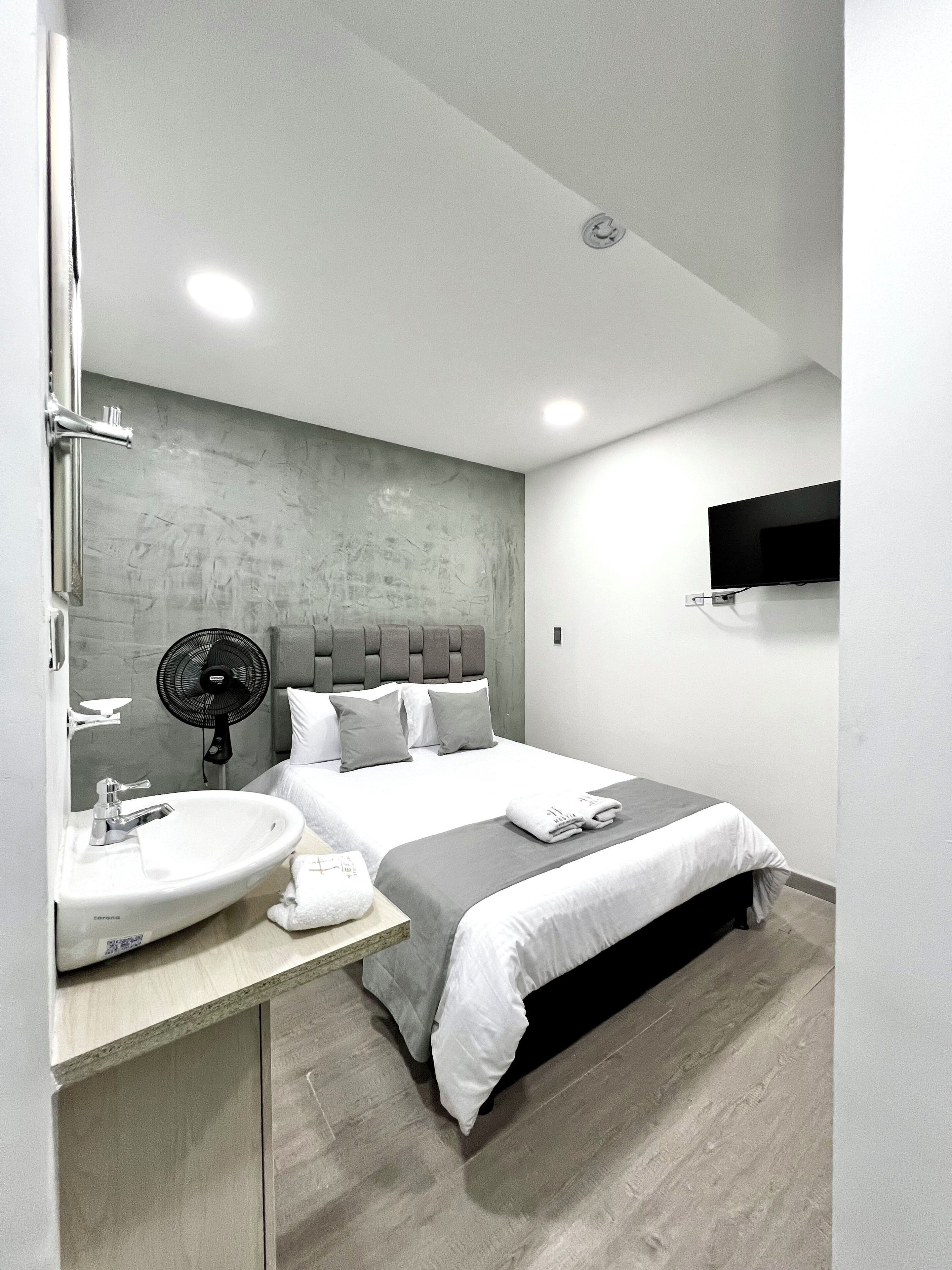 A modern hotel room with a large bed, a bedside table with a sink, a fan, and a flat-screen TV mounted on the wall.