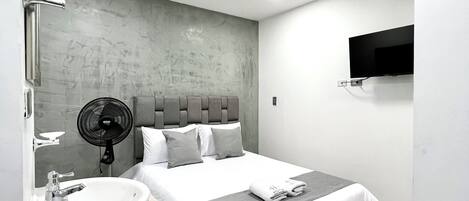 A modern hotel room with a large bed, a bedside table with a sink, a fan, and a flat-screen TV mounted on the wall.