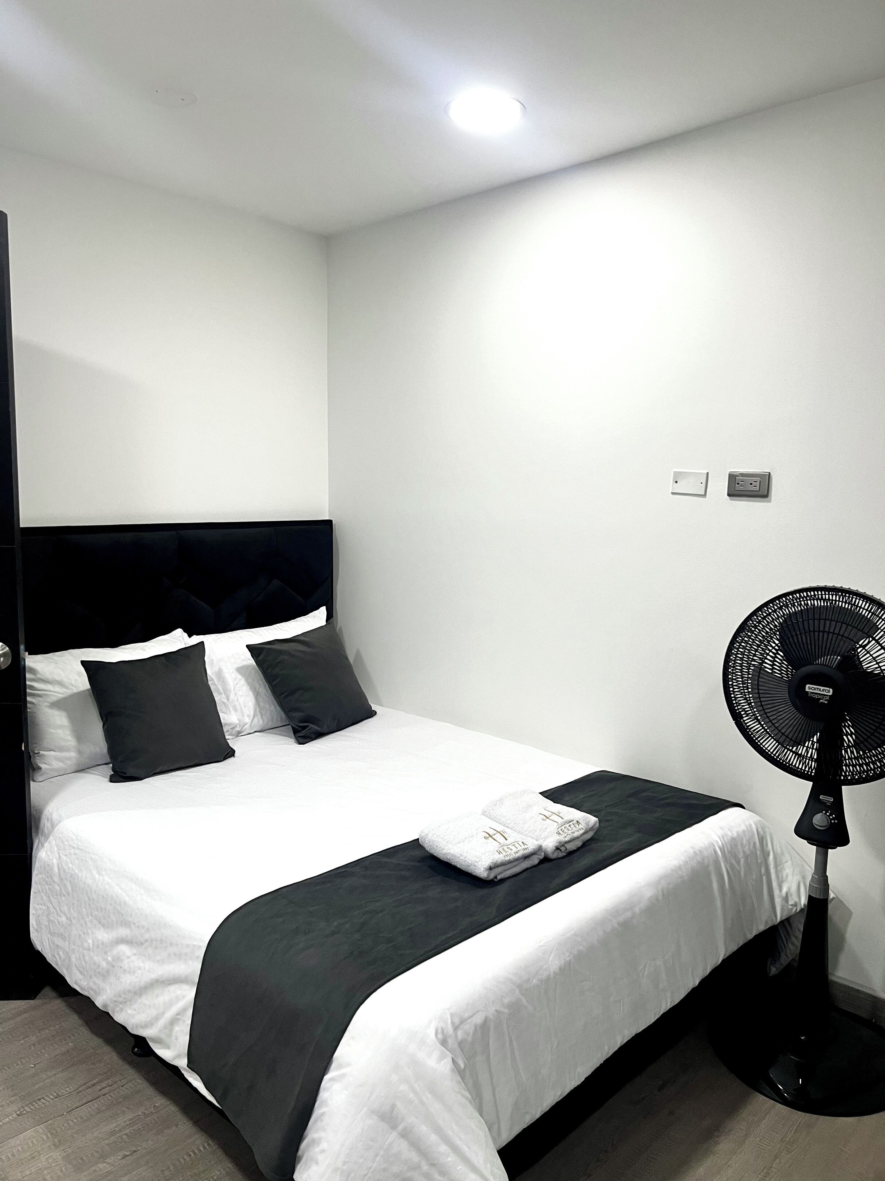 A neatly made bed with white and black bedding, a black fan, and a simple wall with two electrical outlets.