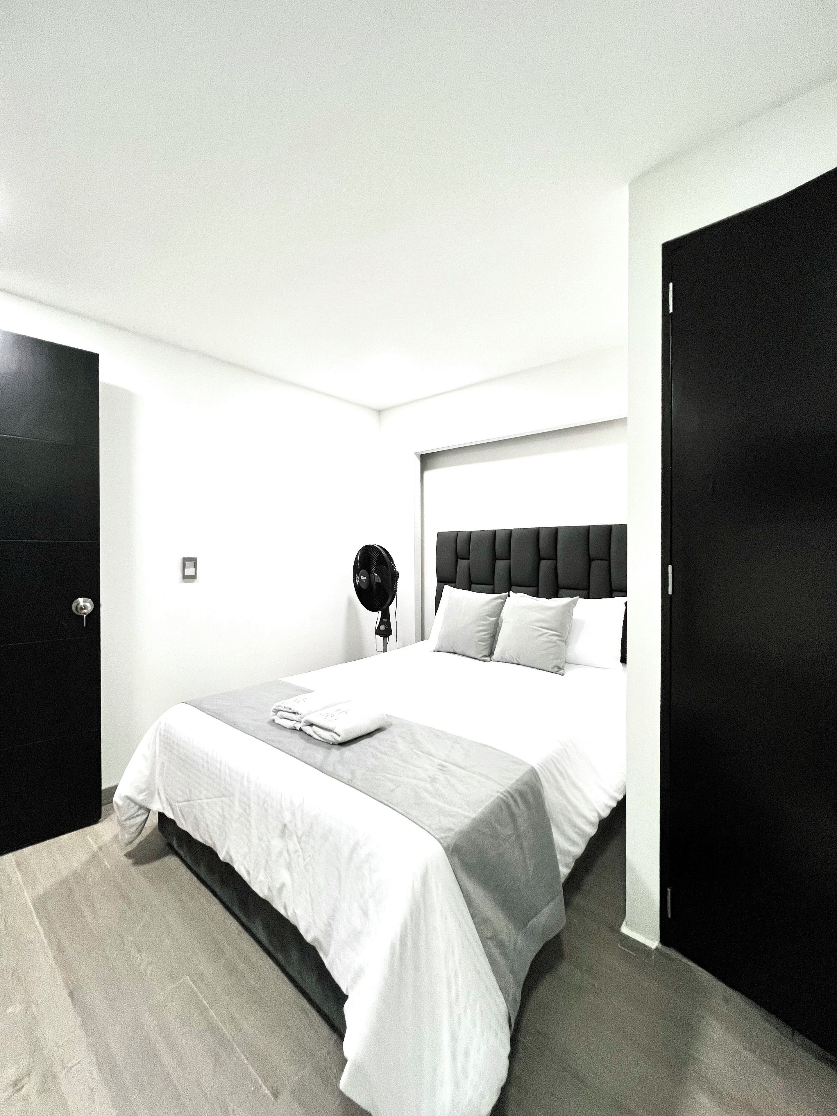 A modern hotel room with a large bed, a bedside fan, and a black wardrobe.